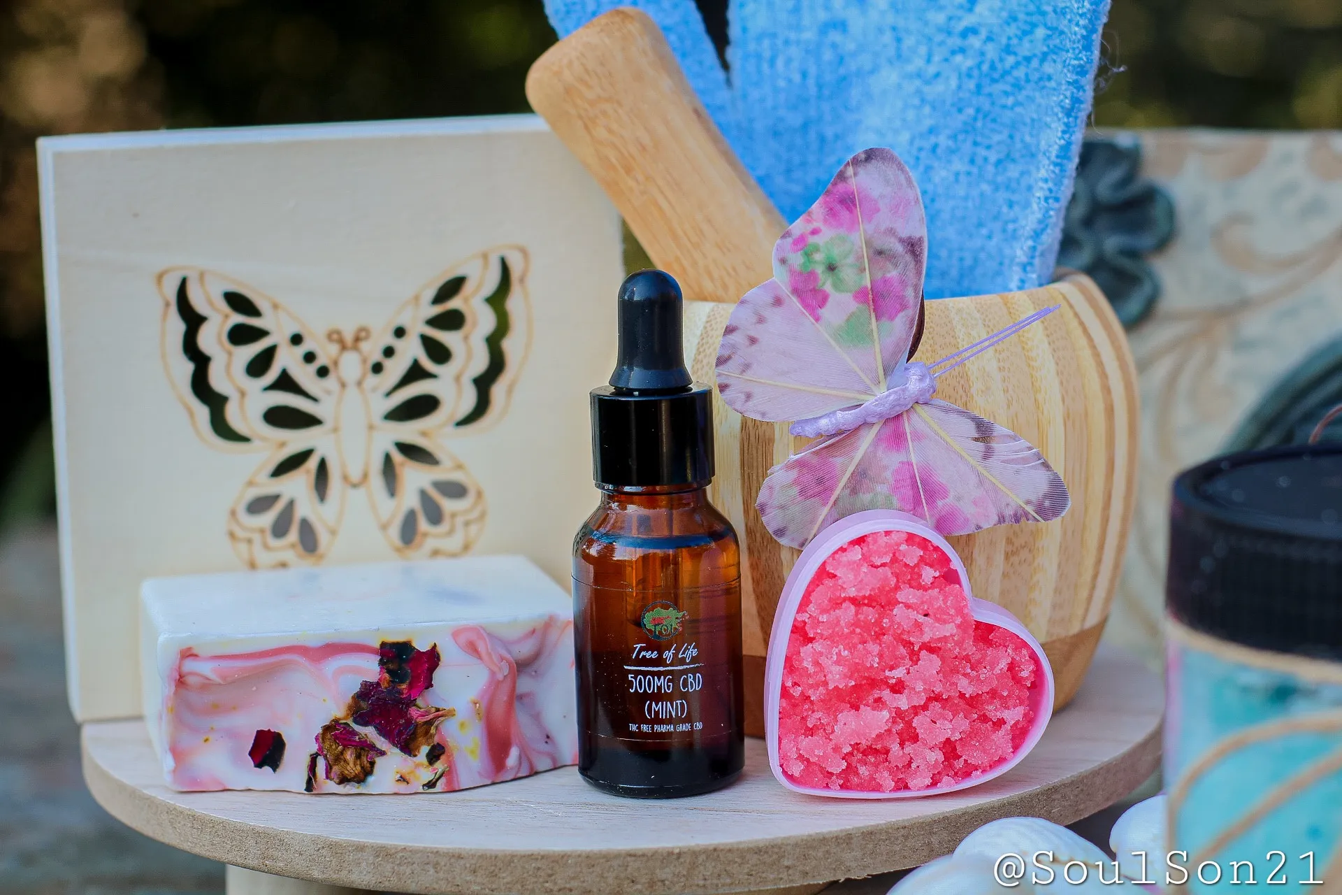 A wooden tray displays a butterfly design, CBD oil, colorful soap, pink bath salts in a heart shape, and an elegant decor arrangement