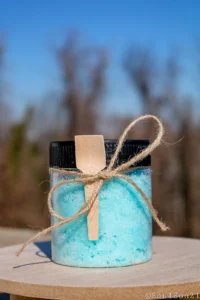 A clear jar filled with blue bath salts, adorned with a wooden spoon and twine, sits on a wooden surface against a blurred background