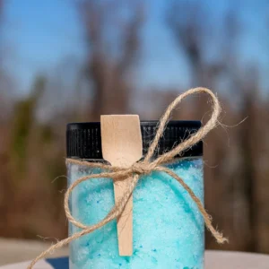 A clear jar filled with blue bath salts, adorned with a wooden spoon and twine, sits on a wooden surface against a blurred background