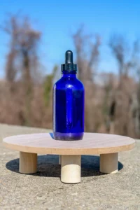 A cobalt blue dropper bottle sits on a circular wooden platform against a clear blue sky with soft, blurred trees in the background