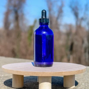 A cobalt blue dropper bottle sits on a circular wooden platform against a clear blue sky with soft, blurred trees in the background