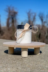 A clear jar filled with white bath salts, adorned with a wooden spoon and twine, sits on a wooden surface against a blurred background