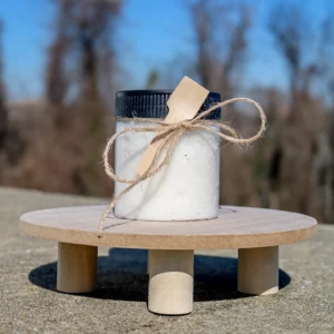 A clear jar filled with white bath salts, adorned with a wooden spoon and twine, sits on a wooden surface against a blurred background