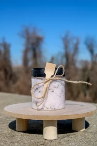 A clear jar filled with lavender bath salts, adorned with a wooden spoon and twine, sits on a wooden surface against a blurred background