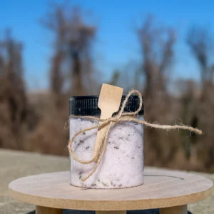 A clear jar filled with lavender bath salts, adorned with a wooden spoon and twine, sits on a wooden surface against a blurred background