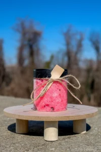 A clear jar filled with pink bath salts, adorned with a wooden spoon and twine, sits on a wooden surface against a blurred background