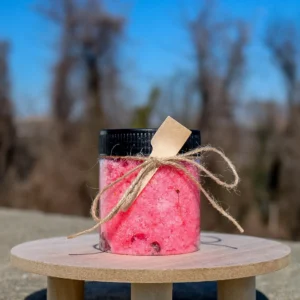 A clear jar filled with pink bath salts, adorned with a wooden spoon and twine, sits on a wooden surface against a blurred background