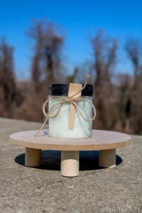 A clear jar filled with green bath salts, adorned with a wooden spoon and twine, sits on a wooden surface against a blurred background