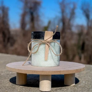 A clear jar filled with green bath salts, adorned with a wooden spoon and twine, sits on a wooden surface against a blurred background