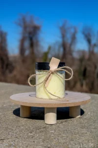 A clear jar filled with yellow bath salts, adorned with a wooden spoon and twine, sits on a wooden surface against a blurred background
