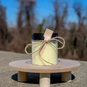 A clear jar filled with yellow bath salts, adorned with a wooden spoon and twine, sits on a wooden surface against a blurred background