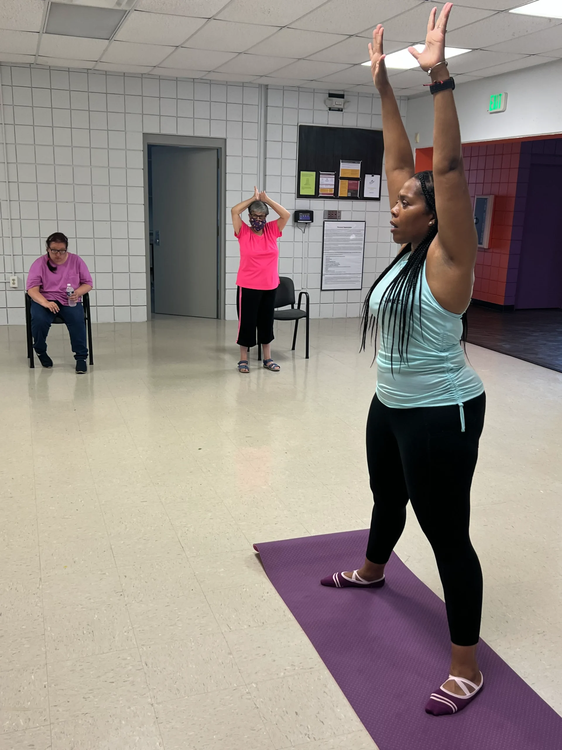 A group exercise class in a bright room, featuring participants engaged in movements with mats and chairs
