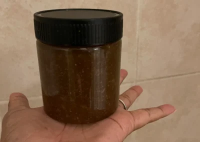 A person holds a small, dark brown jar with a black lid, set against a light-colored tiled background