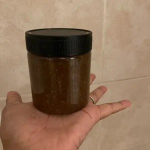 A person holds a small, dark brown jar with a black lid, set against a light-colored tiled background