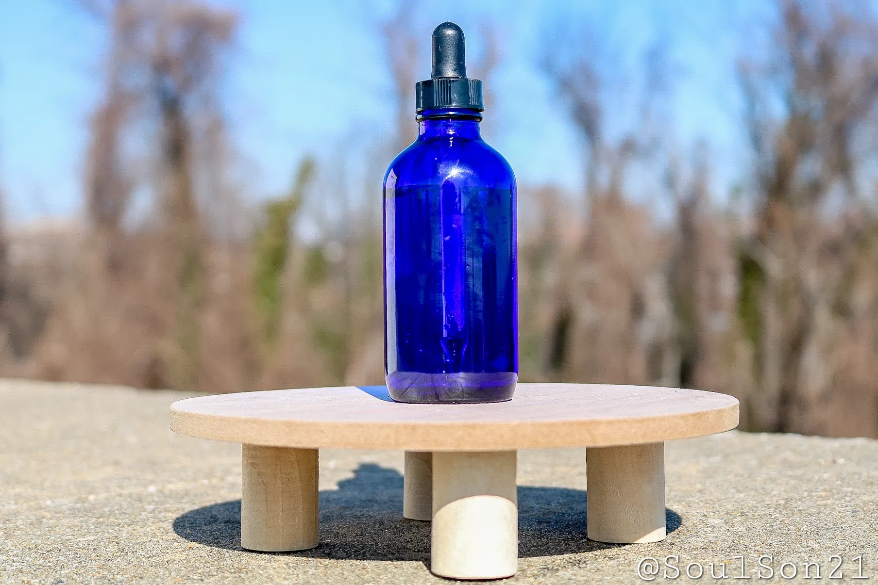 cobalt blue dropper bottle sits on a circular wooden platform against a clear blue sky with soft, blurred trees in the background